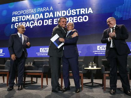O presidente Jair Bolsonaro no palco de um evento promovido pela Confederação Nacional da Indústria (CNI). Ele está abraçando o ministro da Economia, Paulo Guedes, e sendo aplaudido por industriais.