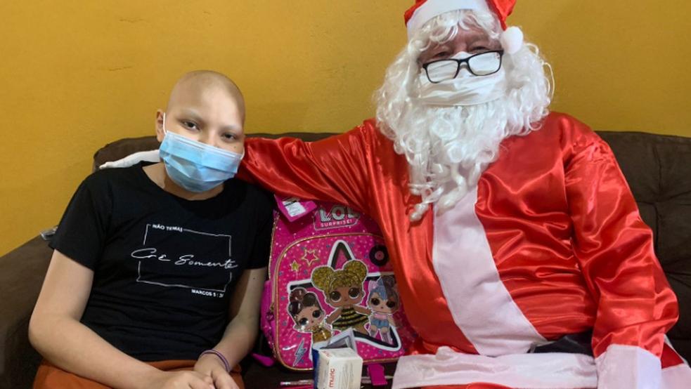 Tailane Alves da Silva sentada em um sofá ao lado do Papai Noel e de seu presente