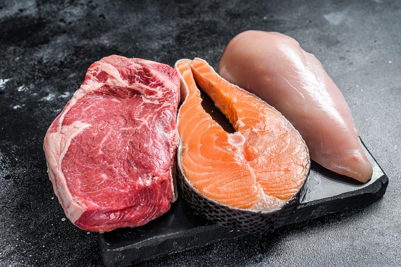 Pedaços de carne bovina, peixe e frango dispostos em um tábua escura sobre um fundo preto