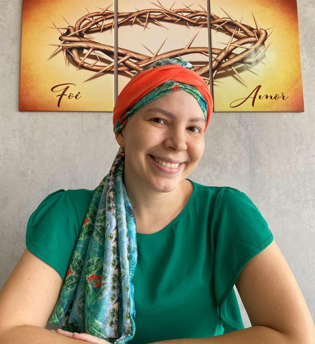 Jornalista Marina Alves posa sorrindo, com blusa verde e lenço colorido na cabeça. Ao fundo, quadro mostra coroa de espinhos e a frase: 