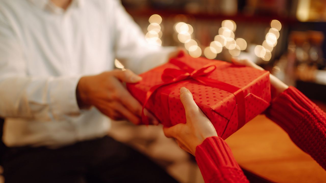 Mãos tocando uma caixa de presente vermelha, com luzes de Natal ao fundo