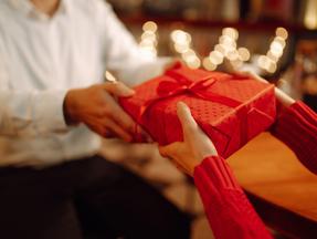 Mãos tocando uma caixa de presente vermelha, com luzes de Natal ao fundo