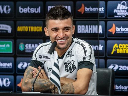 Jogador está sentado, sorrindo, vestindo a camisa do clube em coletiva de imprensa