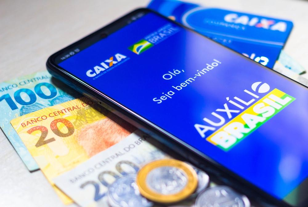 A imagem mostra um celular no aplicativo da Caixa para receber o Auxílio Brasil. O celular está sobre cédulas e moedas.