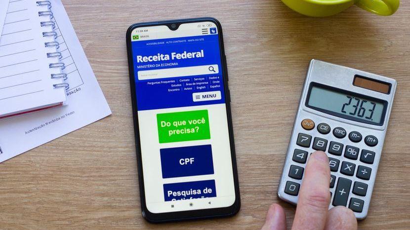 Sobre uma mesa, calculadora, agenda e um celular aberto com um aplicativo da Rceita Federal.