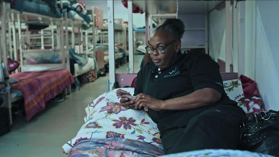 Cena de Onde eu Moro: Mulher negra olha o celular deitada na cama de um albergue