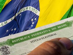 Uma mão segurando um título eleitoral de frente para a bandeira do Brasil.