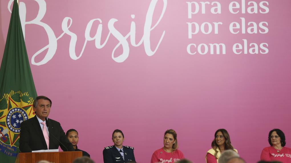 bolsonaro diante de fundo cor de rosa ao lado de quatro mulheres durante solenidade
