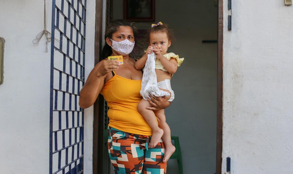 Mulher segura criança no colo com um braço e, com o outro, estende um cartão em que recebe benefício do Governo.