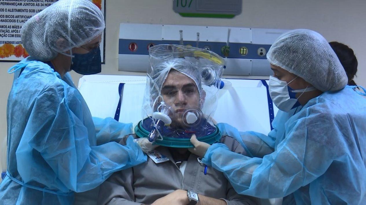 Paciente com Covid-19 utilizando o capacete Elmo. Duas profissionais da saúde sustentam o equipamento.