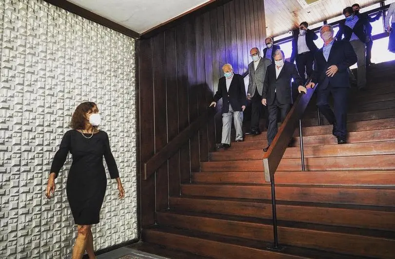 Izolda Cela de vestido preto, sozinha, com o corpo e a cabeça levemente inclinados para trás, olhando um grupo de homens descer as escadas do Palácio da Abolição: o então governador Camilo Santana e os ex-governadores Tasso Jereissati (PSDB), Ciro e Cid Gomes (PDT), Lúcio Alcântara (PL), Chico Aguiar e Gonzaga Mota.