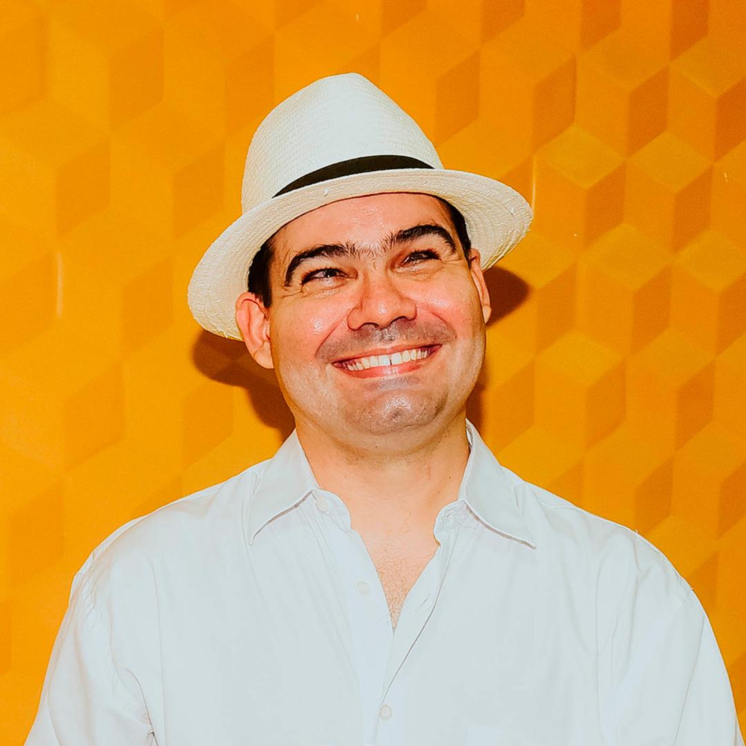 João Bosco Farias, professor cearense cego, sorri, vestindo blusa de botão e chapéu brancos. Ao fundo, uma parede amarela com mosaicos.