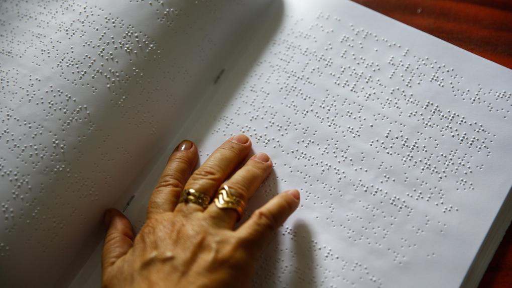 Imagem mostra mão de uma mulher, com anéis nos dois dedos do meio, lendo um texto em braille.
