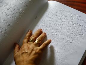Imagem mostra mão de uma mulher, com anéis nos dois dedos do meio, lendo um texto em braille.