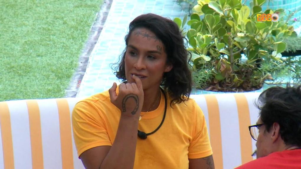 A cantora e participante do BBB, Lina Pereira, está sentada de camisa amarela. Ela está roendo a unha do dedo mindinho.