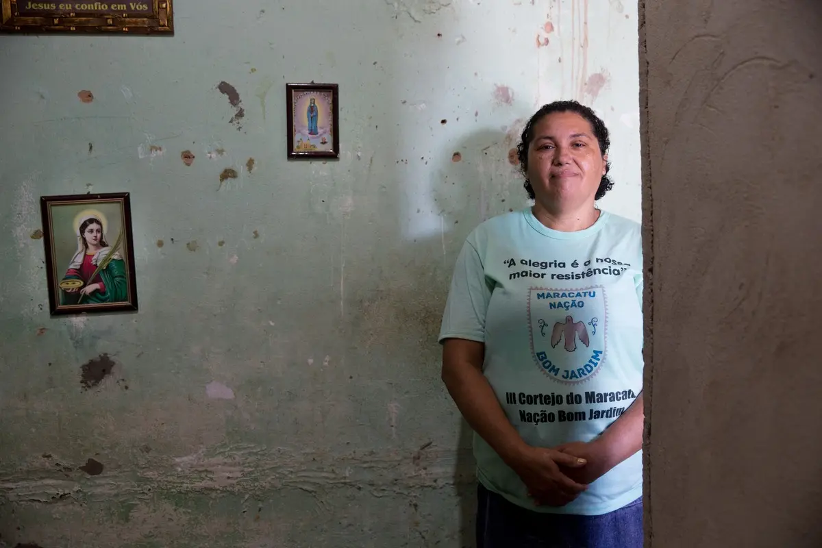 Personagem encostada numa parede verde. Atrás, dois quadros de santos