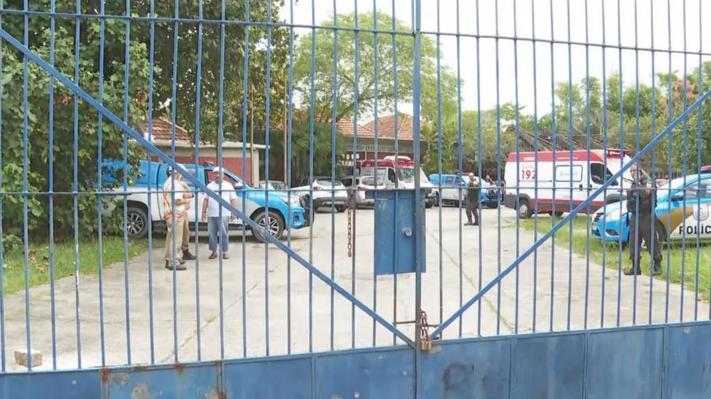 portão azul de escola fechado; no pátio, viaturas e ambulâncias