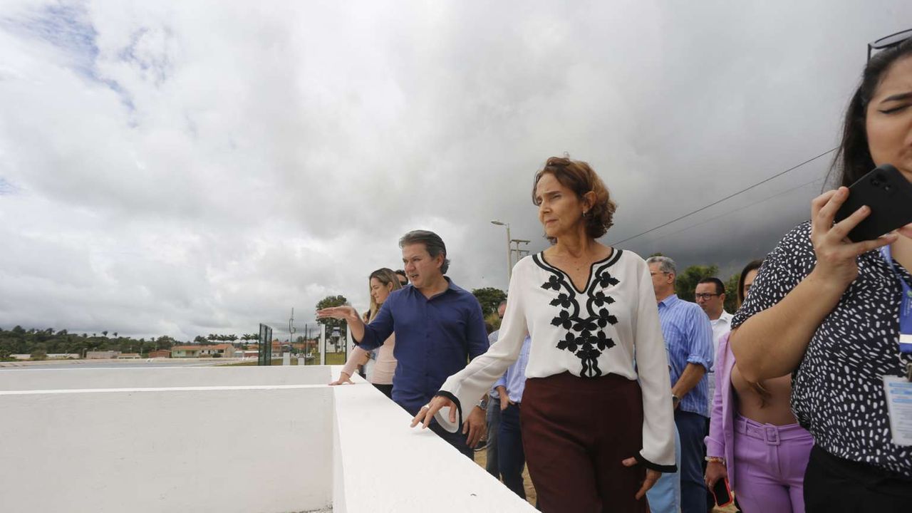 A governadora Izolda Cela aparece em primeiro plano, vestindo uma camisa branca e uma calça marrom, observando um sistema de abastecimento de água em Tianguá. O céu está nublado.