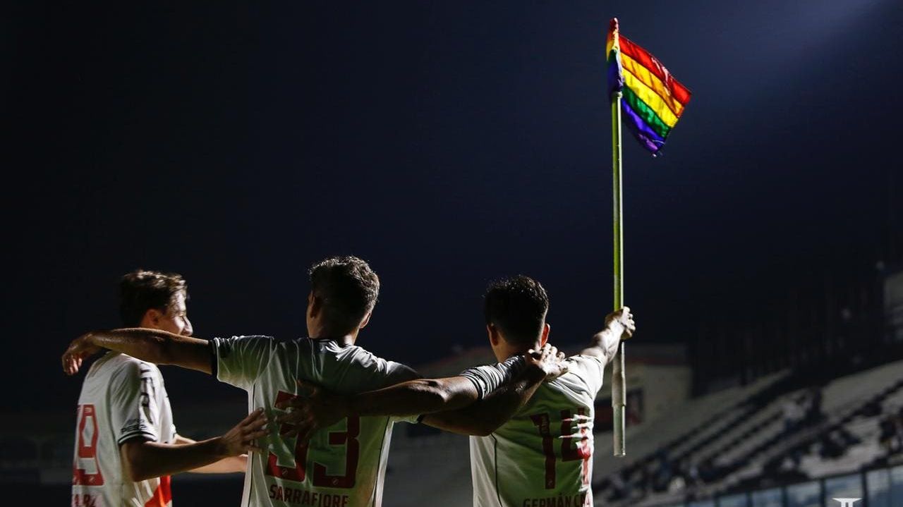 Imagem mostra jogadores abraçados e levantando a bandeira do arco-íris