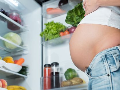 barriga de mulher grávida diante de geladeira aberta com verduras e frutas