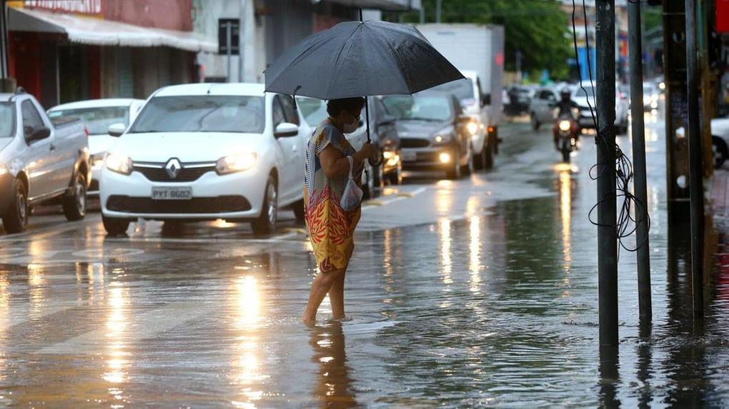 Mulher atravessa poça d'água em rua movimentada de Fortaleza. Ela segura um guarda-chuva preto.