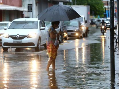 Mulher atravessa poça d'água em rua movimentada de Fortaleza. Ela segura um guarda-chuva preto.