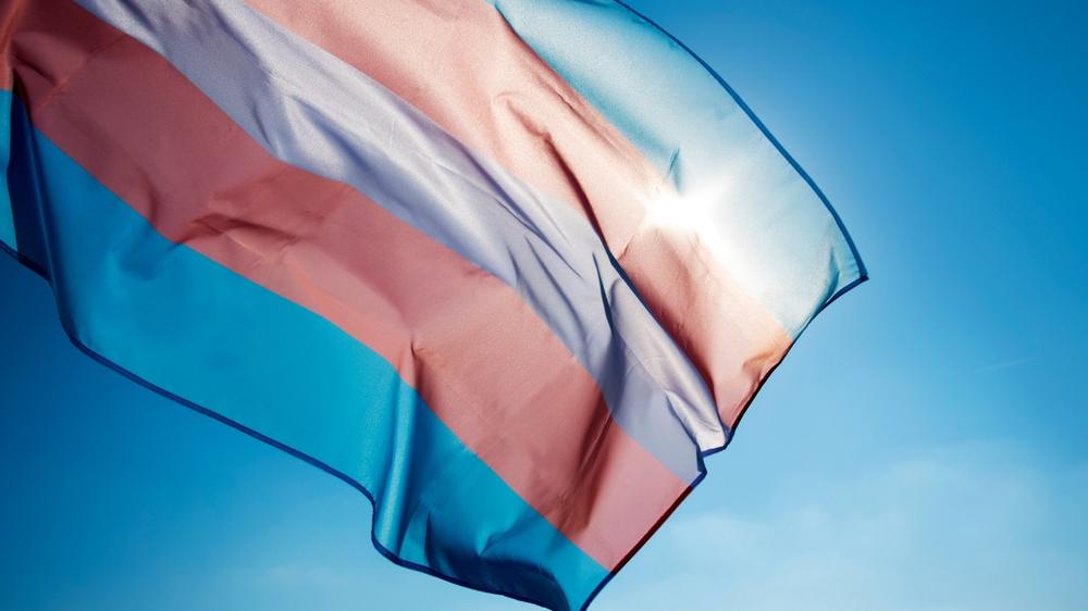 Bandeira do orgulho transgênero acenando no céu azul, movida pelo vento, com o sol ao fundo.