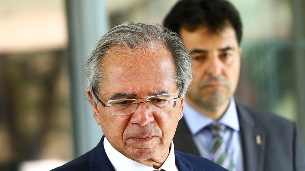 O ministro da Economia, Paulo Guedes, de cabeça baixa. Ele está de terno e gravata verde.
