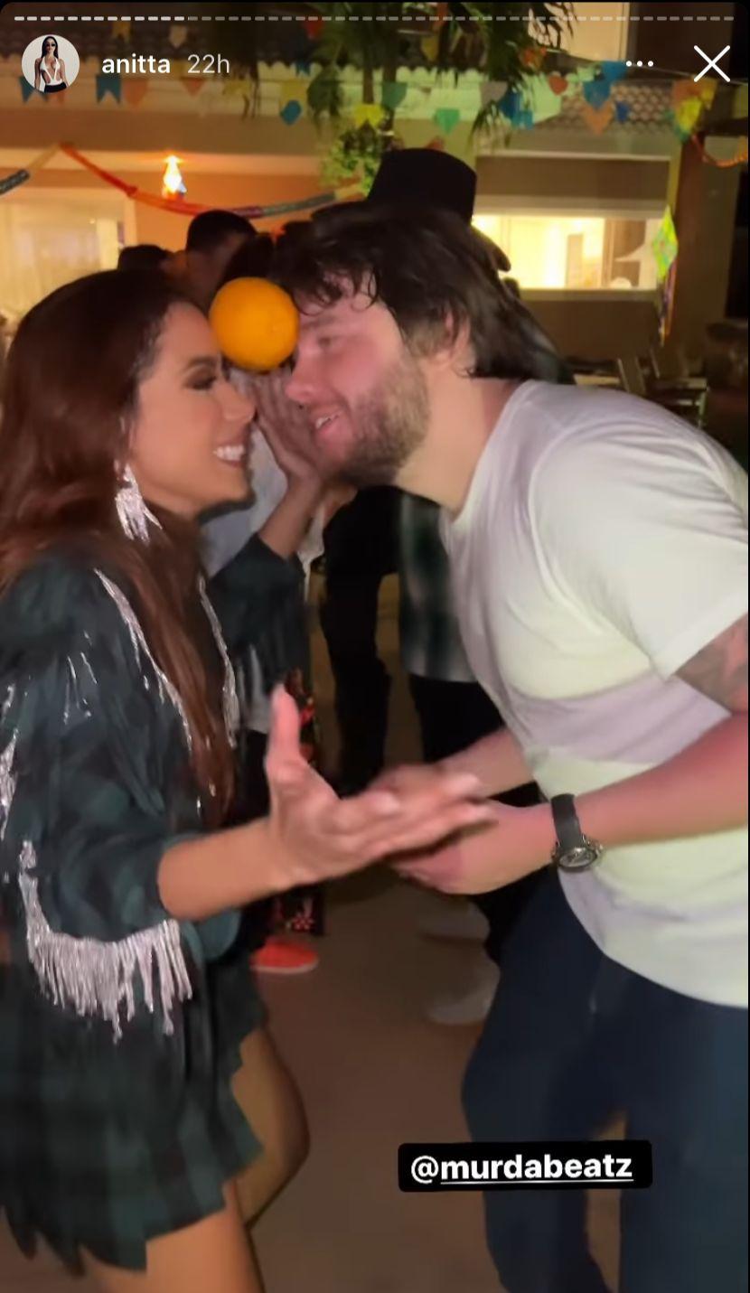 A cantora Anitta e o produtor Murda Beatz fazem a dança da laranja. Eles estão de frente para o outro sustentando uma laranja na testa.
