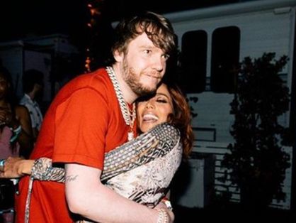 O produtor Murda Beatz está de camisa vermelha abraçando a cantora Anitta, que está de olhos fechados e sorrindo.