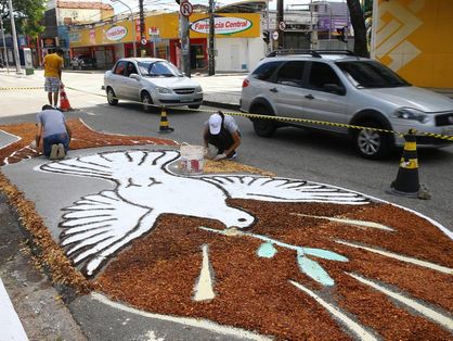 Moradores de Fortaleza confeccionam tapete gigante para o Dia de Corpus Christi. Eles estão em uma avenida da cidade. Carros passam ao fundo da imagem.