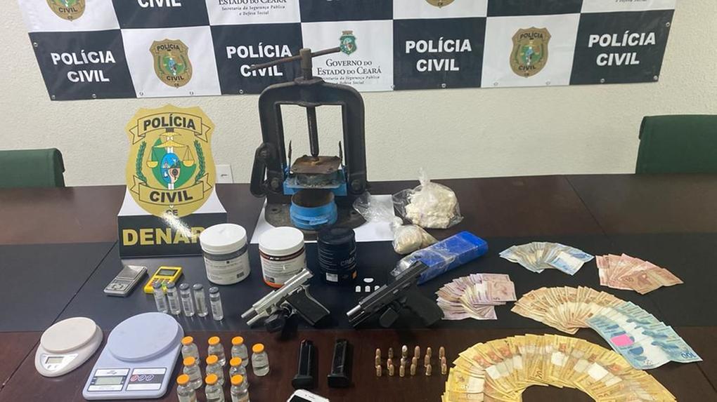 Sobre uma mesa de uma delegacia estão drogas, dinheiro em espécie, munições, revólveres, balanças de precisão e outros apetrechos utilizados para o tráfico de drogas.