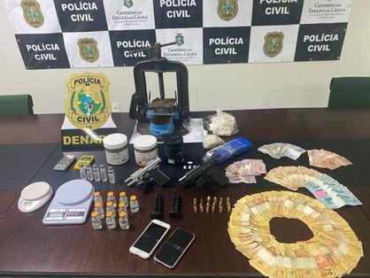 Sobre uma mesa de uma delegacia estão drogas, dinheiro em espécie, munições, revólveres, balanças de precisão e outros apetrechos utilizados para o tráfico de drogas.