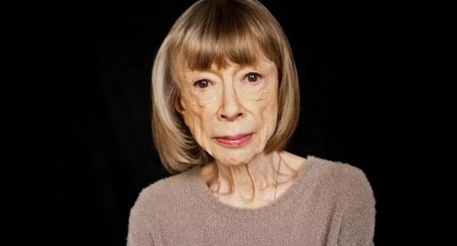 A escritora Joan Didion loira, de cabelo curto cortado chanel. Ela usa uma blusa marrom e está séria. Fundo da imagem é preto.