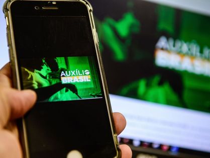 Mão segura celular com imagem de banner do Auxílio Brasil aberta na tela