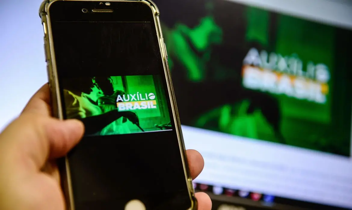 Mão segura celular com imagem de banner do Auxílio Brasil aberta na tela
