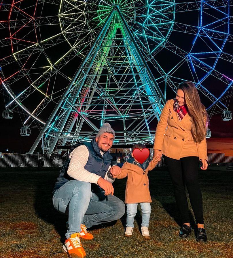 Família em frente a uma roda gigante. O rosto de Santiago é coberto por um emoji de coração.