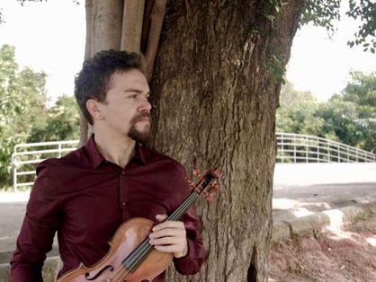 O violinista Lucas Raulino segura um violino em frente a uma árvore. Ele está em um parque e usa camisa vermelha de botão.