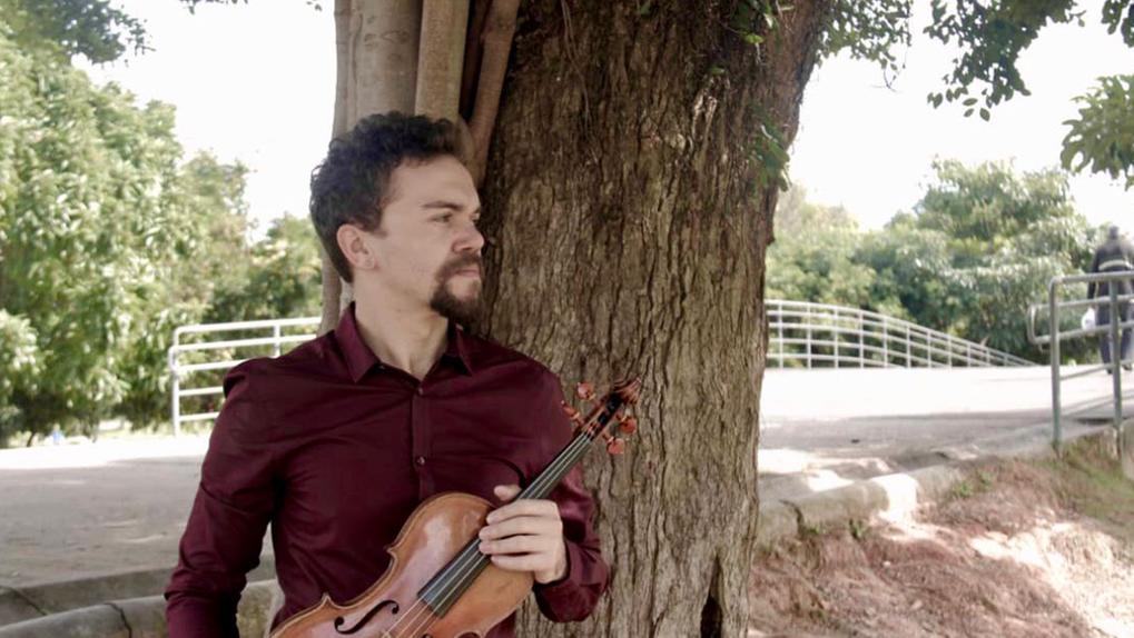 O violinista Lucas Raulino segura um violino em frente a uma árvore. Ele está em um parque e usa camisa vermelha de botão.