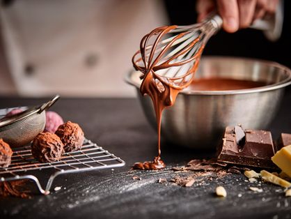 Padeiro preparando bombons de chocolate, mexendo o chocolate derretido com um batedor pingando no balcão abaixo