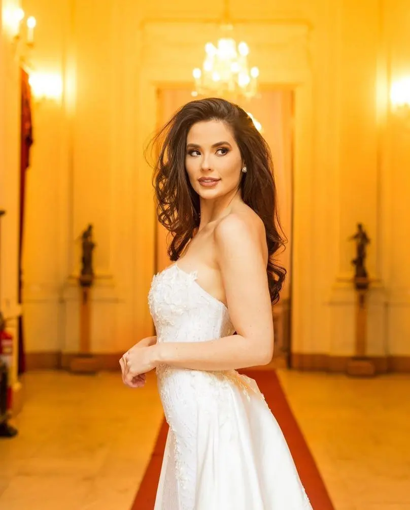 A modelo Alina Furtado está em um vestido branco. Seu cabelo é longo e castanho escuro.