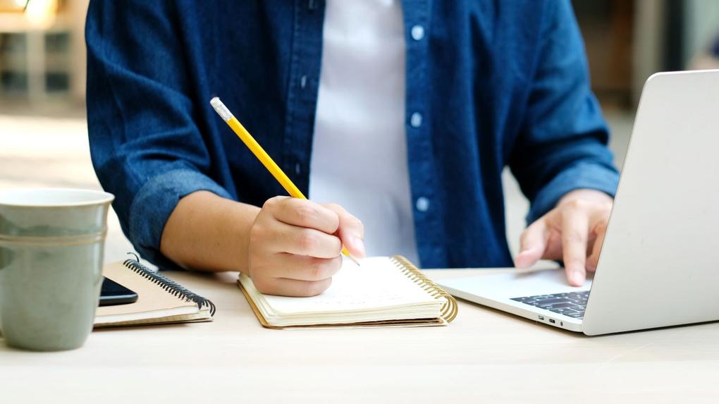 pessoa com camisa azul escreve em caderno enquanto mexe em notebook