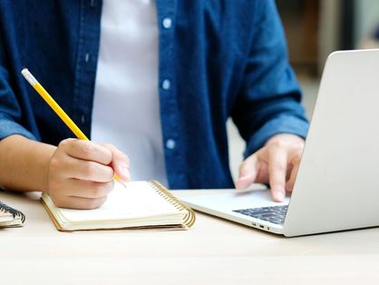 pessoa com camisa azul escreve em caderno enquanto mexe em notebook