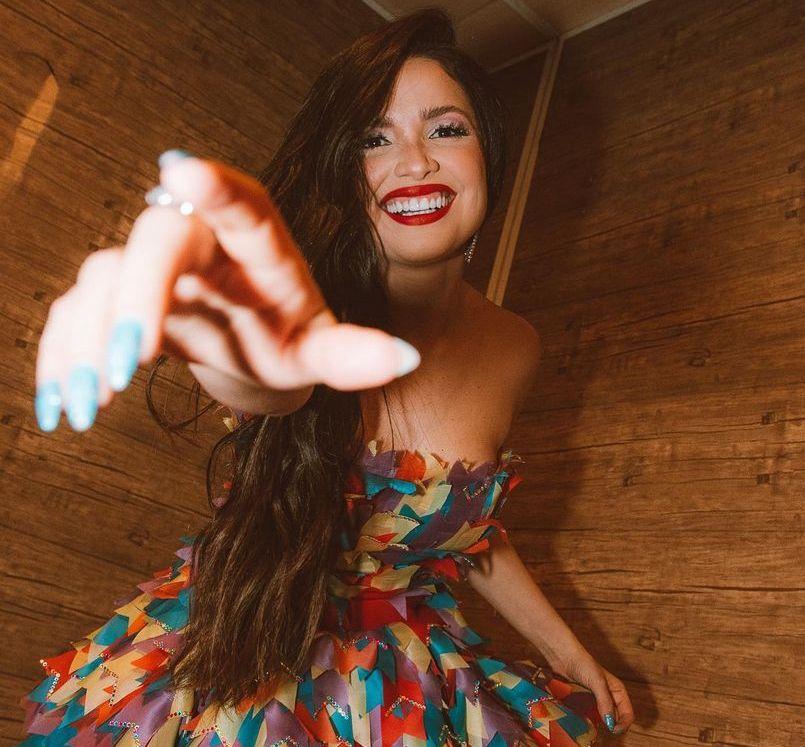 A cantora Juliette está sorrindo e de batom vermelho. Ela está com a mão estendida como se fosse pegar a câmera.