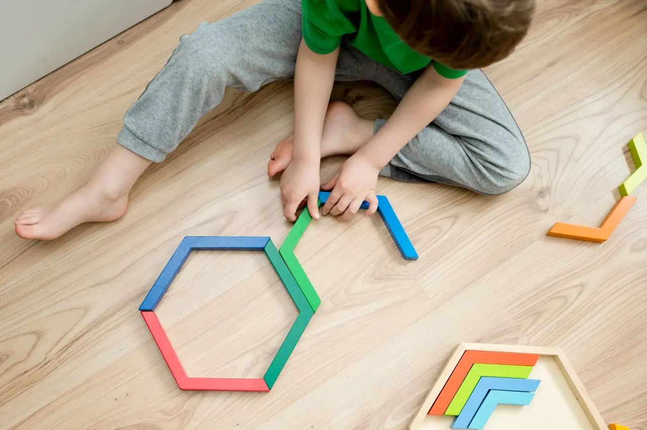 criança sentada em chão de madeira com calça cinza e camiseta verde brincando com brinquedos educativos