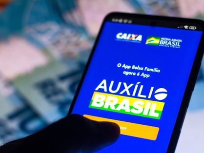Mão segura um celular aberto no aplicativo do Auxílio Brasil. Ao fundo, cédulas de 100 reais.