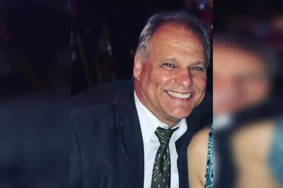 Foto da vítima sorrindo. É uma home grisalho, de olhos claros que veste um blazer preto, camisa branca e gravata.