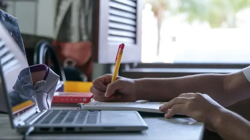 Criança escrevendo em caderno ao lado de notebook.