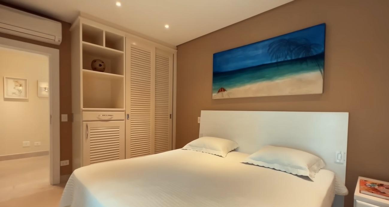 Um dos quartos da mansão de Hebe Camargo. A pintura é marrom e bege. Há uma pintura de praia em cima da cama, que tem colcha branca.
