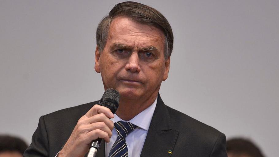 Jair Bolsonaro segura microfone. Ele está sério e usa terno preto, camisa branca e gravata.